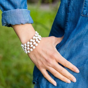 May include: A white beaded bracelet with gold accents. The bracelet is layered and worn on a person's wrist.