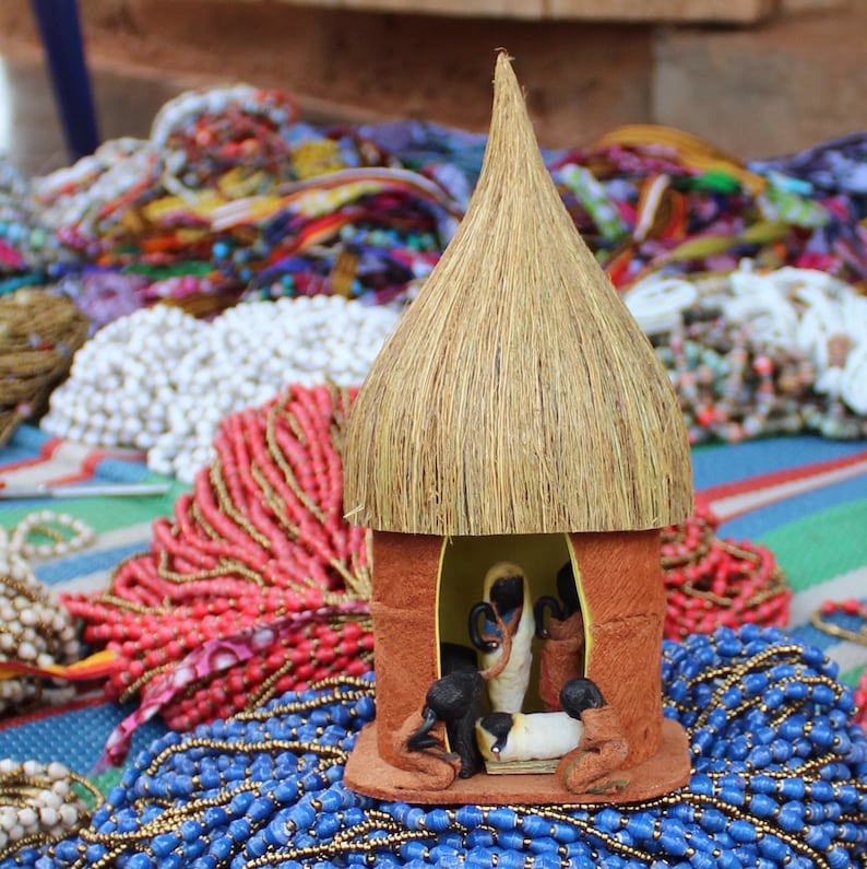 May include: A small thatched hut figurine with a nativity scene inside. The hut is surrounded by colorful beaded necklaces.