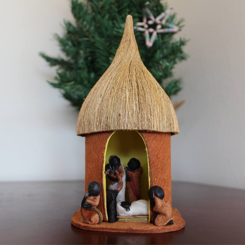 May include: A small, brown, thatched-roof hut with a yellow interior. Inside, there are figures of people and a baby lying in a manger. This is a traditional nativity scene.