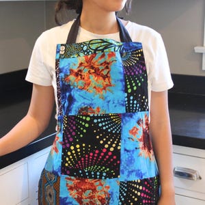 May include: A colorful patchwork apron with a vibrant design. The apron features a mix of blue, orange, and black patterned fabric, with a tie-around neck and waist. The apron is worn over a white t-shirt.