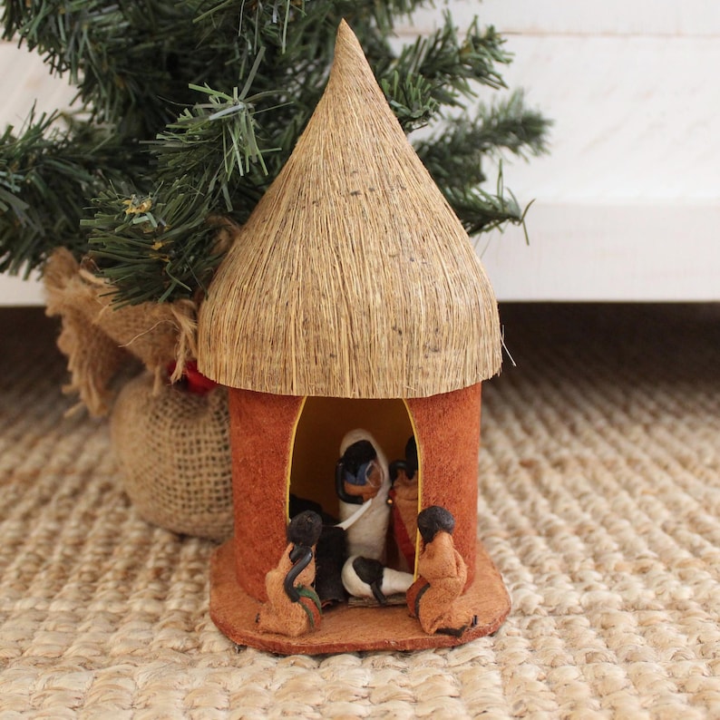 May include: A small, hand-crafted hut made of straw and fabric with a thatched roof. Inside the hut are four small figures, two of which are holding a small, white object. The hut is sitting on a woven rug.