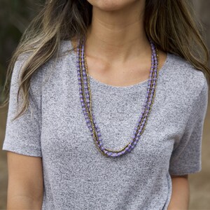 May include: A layered necklace with purple and gold beads. The necklace is worn by a person with long brown hair and a grey sweater.
