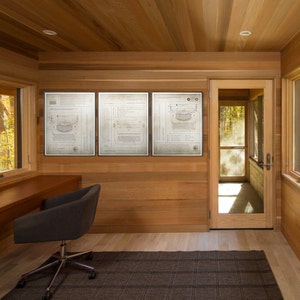 May include: Three framed black and white blueprints of a vintage typewriter hang on a wood paneled wall in a home office. A dark brown desk with a black office chair sits in front of the wall. A glass door leads to a screened porch.