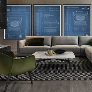 May include: Three blueprint style posters of vintage typewriters, hanging on a wall above a gray sectional sofa and a green velvet armchair. The posters feature the IBM Selectric I, Underwood Portable Royal Standard, and Underwood 1930s Portable Royal Standard.