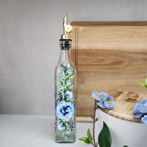Può includere: Una bottiglia per olio in vetro trasparente con un beccuccio in metallo dorato. La bottiglia presenta un disegno dipinto a mano di fiori blu e foglie verdi che corrono verticalmente lungo la parte anteriore. Il vetro è quadrato con bordi arrotondati.