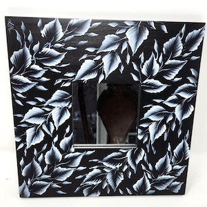 May include: A black wood framed mirror with a white leaf design painted on the frame. The mirror is rectangular and has a beveled edge.