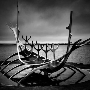 May include: A black and white photo of a large metal sculpture of a ship with a long, pointed bow and a series of curved, branching arms. The sculpture is set against a cloudy sky and a body of water.