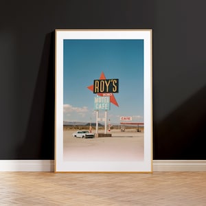 May include: Framed print featuring a vintage roadside scene. The image showcases a retro sign for "Roy's Motel Cafe" against a clear blue sky. A classic white car is parked nearby, with a gas station and desert landscape in the background.