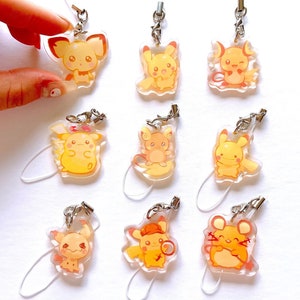 Puede incluir: Colección de nueve colgantes de personajes de dibujos animados amarillos y naranjas, con cierres plateados y correas transparentes. Los colgantes son sostenidos por una mano sobre un fondo blanco. Cada colgante presenta una pose diferente.