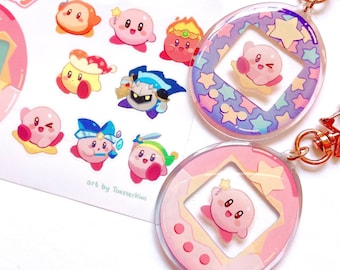 May include: Two pink and white Tamagotchi keychains with a pink and white sticker sheet featuring Kirby characters. The keychains feature a pink and white Tamagotchi with a clear window and a pink Kirby character inside. The sticker sheet features a pink Tamagotchi with a clear window and a pink Kirby character inside, as well as other Kirby characters.