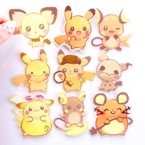 Puede incluir: Una colección de nueve pegatinas de personajes de dibujos animados en varias poses. Las pegatinas presentan personajes amarillos, tostados y marrones con contornos negros. Una mano sostiene una pegatina. Las pegatinas están sobre un fondo blanco.