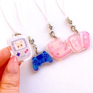 May include: Clear acrylic charms featuring gaming-themed designs. One charm depicts a retro handheld console with a cute hamster, while others showcase a pink console, controllers, and a blue gamepad. Each charm has a white lanyard.