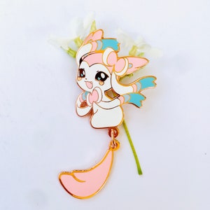 May include: A gold-colored enamel pin featuring a cartoon character with pink, blue, and white details. The character has a long, pink tail that dangles from the pin.