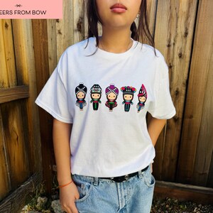 May include: White t-shirt featuring a row of six cartoon figures in traditional outfits. Each figure has a unique hat and outfit design. The text "CHEERS FROM BOW" is in a pink rectangle at the top.