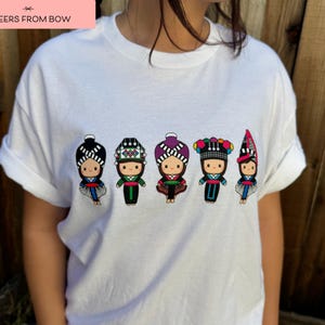 May include: White t-shirt featuring a row of five cartoon figures in traditional ethnic clothing. Each figure has a unique hat and outfit design, with details in black, red, green, and purple. The text "CHEERS FROM BOW" is in a pink rectangle in the top left corner.