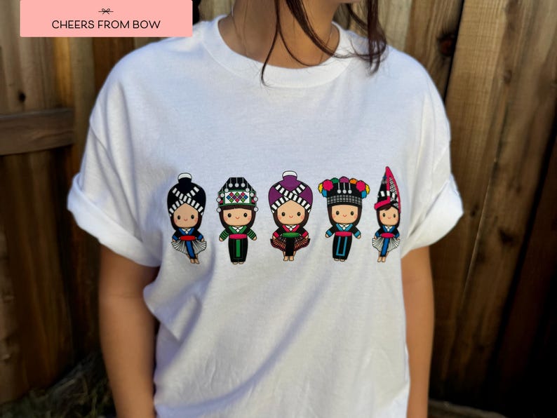 May include: White t-shirt featuring a row of five cartoon figures in traditional ethnic clothing. Each figure has a unique hat and outfit design, with details in black, red, green, and purple. The text "CHEERS FROM BOW" is in a pink rectangle in the top left corner.
