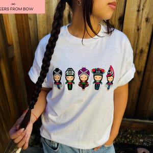 May include: White t-shirt featuring a colorful graphic of five stylized figures in traditional clothing. The figures have detailed headwear and outfits in various colors. The text "CHEERS FROM BOW" is in a pink rectangle in the upper left corner.