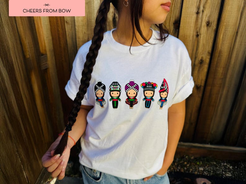 May include: White t-shirt featuring a colorful graphic of five stylized figures in traditional clothing. The figures have detailed headwear and outfits in various colors. The text "CHEERS FROM BOW" is in a pink rectangle in the upper left corner.