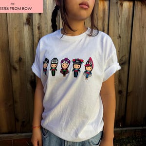 May include: White t-shirt featuring a colorful embroidered design of five figures in traditional clothing. The figures are wearing hats and dresses in various colors. The text "CHEERS FROM BOW" is in a pink rectangle.