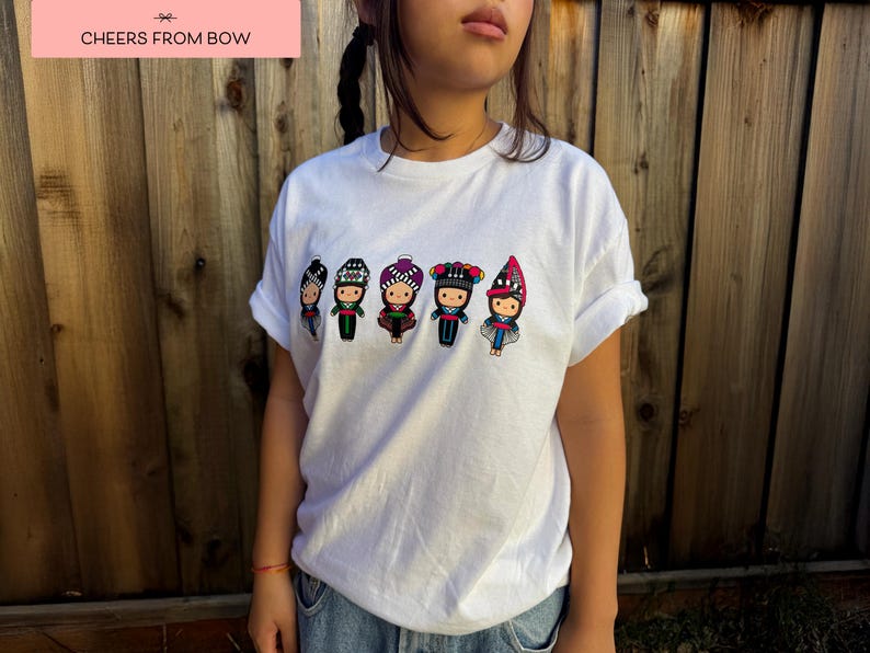 May include: White t-shirt featuring a colorful embroidered design of five figures in traditional clothing. The figures are wearing hats and dresses in various colors. The text "CHEERS FROM BOW" is in a pink rectangle.