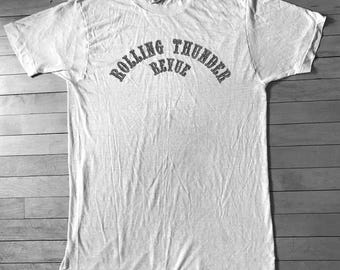 May include: A white t-shirt with the text "Rolling Thunder Revue" printed in black on the front.