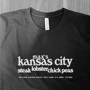May include: Black t-shirt with white text. The text reads "max's kansas city steak lobster chick peas" with an address and phone number below. The shirt is a Bella + Canvas brand.