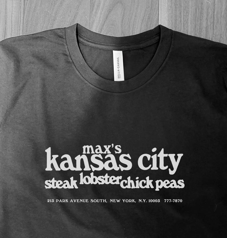 May include: Black t-shirt with white text. The text reads "max's kansas city steak lobster chick peas" with an address and phone number below. The shirt is a Bella + Canvas brand.