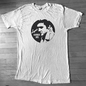 Może przedstawiać: Biały t-shirt z czarno-białym nadrukiem Elvisa Presleya. Nadruk jest w kształcie koła z tekstem "Lupo's Heartbreak Hotel 3777 Westminster, Prov. RI."