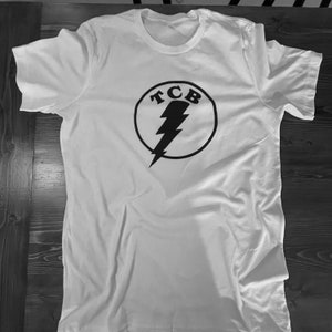 Puede incluir: Camiseta blanca con un logotipo circular negro que presenta un rayo y las letras "TCB".