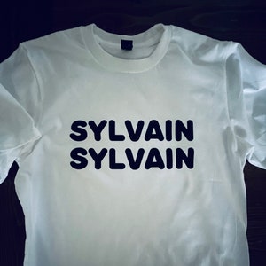 May include: White t-shirt with the word "SYLVAIN" printed twice in black, bold letters.