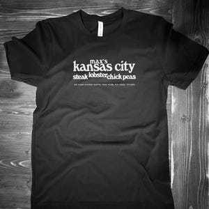 May include: Black t-shirt with white text. The text reads "max's kansas city steak lobster chick peas" with an address and phone number below. The shirt is made of a soft material.