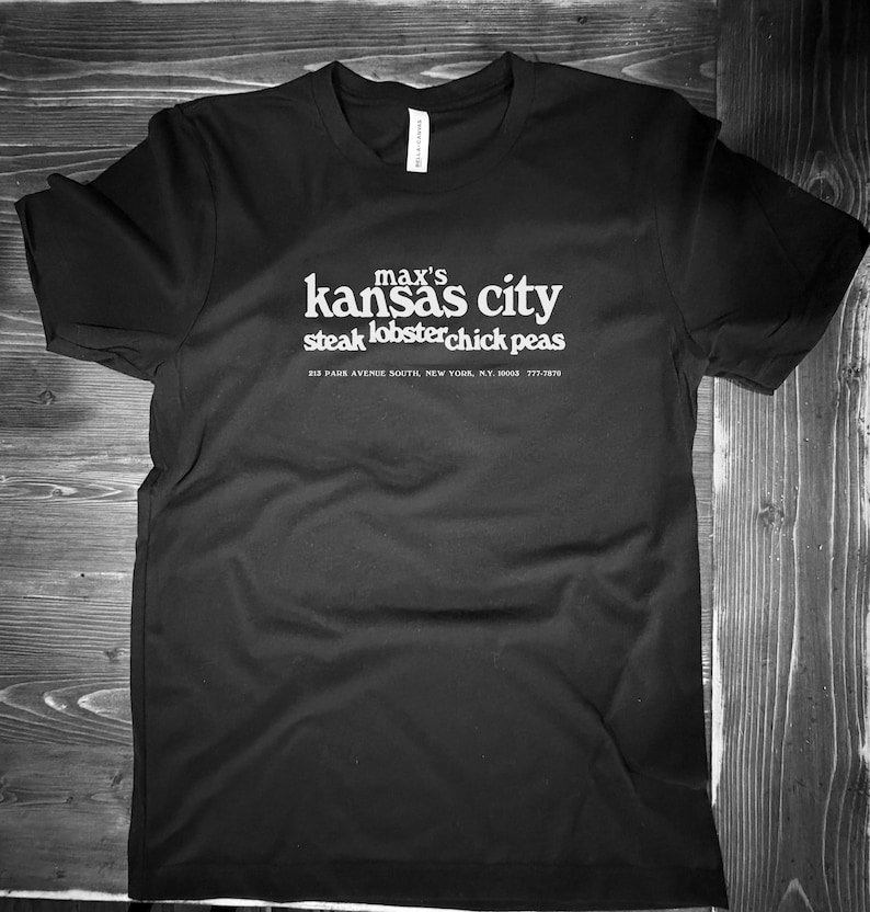 May include: Black t-shirt with white text. The text reads "max's kansas city steak lobster chick peas" with an address and phone number below. The shirt is made of a soft material.