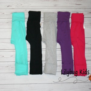 May include: Six pairs of children's leggings in different colors: turquoise, black, gray, purple, and pink. The leggings are folded and laid out on a white wooden surface. The text "Lilybug Kids" is visible in the lower right corner of the image.