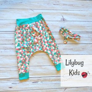 Op de afbeelding: Een harem broek met geometrische print in teal, roze en wit met een teal tailleband. Een bijpassend teal, roze en wit hoofdband is inbegrepen. De tekst "Lilybug Kids" is zichtbaar in de afbeelding.