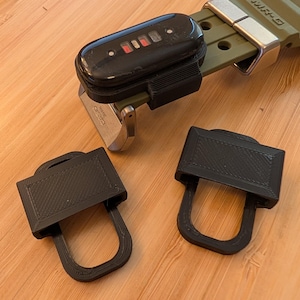 Puede incluir: Un accesorio de correa de reloj de pl&aacute;stico negro impreso en 3D unido a una correa de reloj verde oliva con un rastreador de actividad f&iacute;sica negro. Dos accesorios de pl&aacute;stico negro id&eacute;nticos impresos en 3D se colocan por separado.