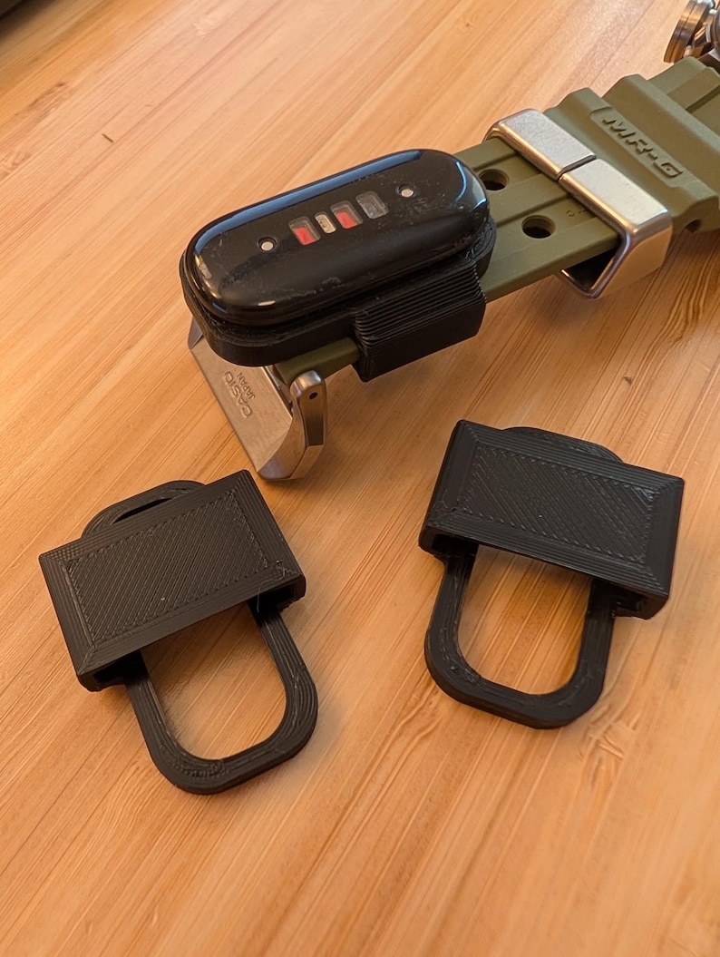 Puede incluir: Un accesorio de correa de reloj de pl&aacute;stico negro impreso en 3D unido a una correa de reloj verde oliva con un rastreador de actividad f&iacute;sica negro. Dos accesorios de pl&aacute;stico negro id&eacute;nticos impresos en 3D se colocan por separado.