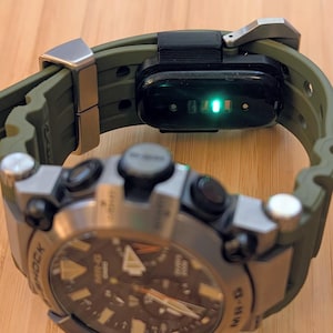 Puede incluir: Un reloj Casio G-Shock MR-G con correa de caucho verde oliva oscuro y caja de metal plateado. En la correa hay un m&oacute;dulo de seguimiento de actividad f&iacute;sico negro con una luz de sensor verde brillante visible en la parte inferior.