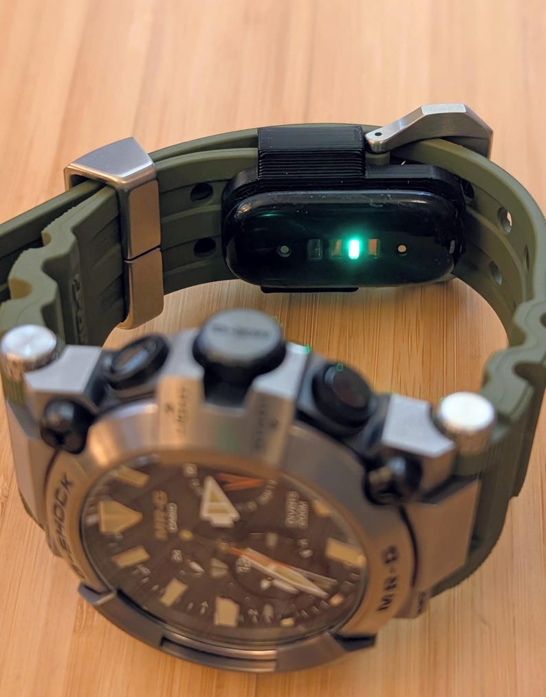 Puede incluir: Un reloj Casio G-Shock MR-G con correa de caucho verde oliva oscuro y caja de metal plateado. En la correa hay un m&oacute;dulo de seguimiento de actividad f&iacute;sico negro con una luz de sensor verde brillante visible en la parte inferior.