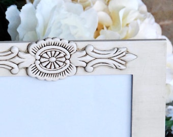 May include: A white wooden picture frame with ornate carvings around the edge. The frame is sitting on a white lace tablecloth.