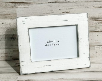 May include: A rectangular white picture frame with distressed edges, displaying the text "jabella designs" in a simple font. The frame is angled slightly, set against a light-colored, wood-grain background.