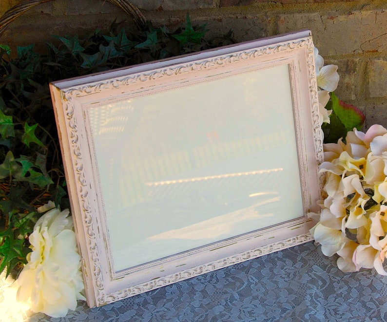 Puede incluir: Un marco de fotos blanco con un acabado rosa desgastado y detalles ornamentados. El marco est&aacute; vac&iacute;o y listo para una foto o una obra de arte.