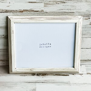 May include: White wood picture frame with a white mat and the text "jabella designs" printed in black.