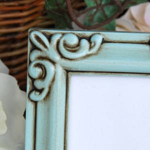 May include: A light blue picture frame with ornate detailing and a distressed finish. The frame has a white mount and is ready to display a photo or artwork.