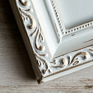 May include: Close-up of a white picture frame with ornate gold detailing. The frame has a distressed finish, with visible wear and tear. The design features intricate scrollwork and a beaded inner edge, creating a vintage aesthetic.