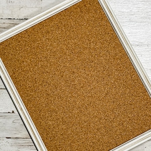 May include: A cork board with a white frame. The cork board is a light brown color and has a textured surface.
