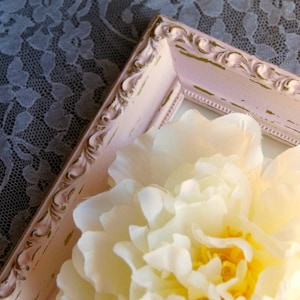 Puede incluir: Una flor blanca con un centro amarillo se muestra en un marco de imagen adornado de color rosa y dorado. El marco tiene un acabado desgastado y est&aacute; sobre un mantel de encaje.