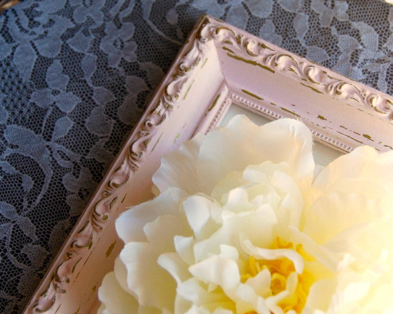 Puede incluir: Una flor blanca con un centro amarillo se muestra en un marco de imagen adornado de color rosa y dorado. El marco tiene un acabado desgastado y est&aacute; sobre un mantel de encaje.