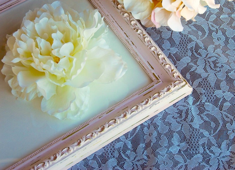 Puede incluir: Una flor de peon&iacute;a blanca prensada y enmarcada en un marco rosa desgastado. El marco est&aacute; sobre un mantel de encaje blanco.