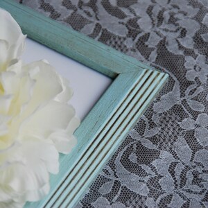 May include: A close-up of a light blue picture frame with a distressed finish. A large, creamy white peony flower is partially visible, along with a white lace fabric background.