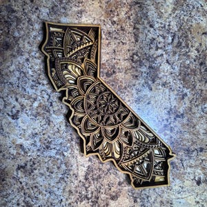 May include: A wooden cutout of the state of California with a detailed mandala design. The mandala is intricate and features a variety of geometric shapes and patterns.
